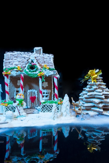 Фото Большой заснеженный самодельный пряничный домик, пряничная елка и снеговик из сахарной мастики на темном фоне. мокап для сезонных предложений и праздничной открытки