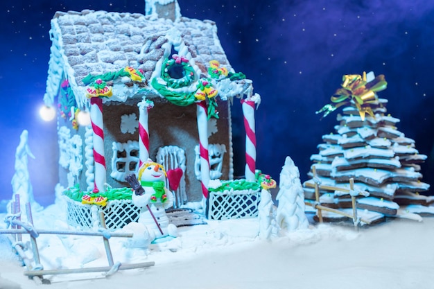 Grande casa di pan di zenzero fatta in casa coperta di neve, albero di natale di pan di zenzero e un pupazzo di neve di mastice di zucchero di notte. mockup per offerte stagionali e cartolina delle vacanze