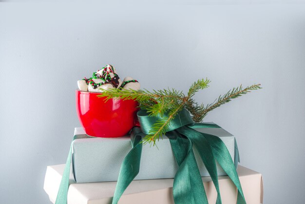 Big stack of Christmas gifts with hot chocolate, stay on it like pedestal, cozy pastel colored craft paper gift boxes with green festive Xmas ribbon, cup of cocoa with marshmallow, copy space