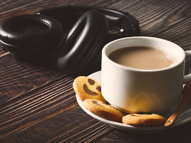 사진 나무 테이블에 행복 한 미소와 함께 커피, 검은 헤드폰 및 노란색 쿠키와 큰 흰색 컵. 확대보기. 행복한 아침과 작업 휴식 개념입니다.