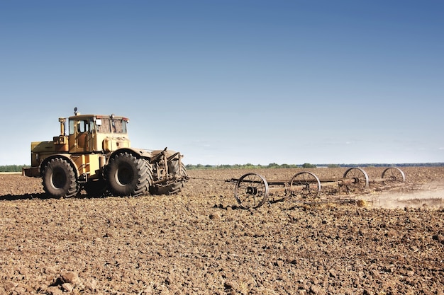 Big yellow tractor equipped with harrow working on the field.