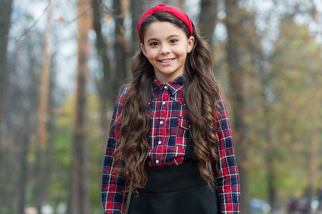 Biggest Hair Accessory Trends. Adorable little girl checkered shirt wear red headband. Fashion trend. Accessories. Fancy child nature background. Padded headband. Smiling girl wear knotted headband.