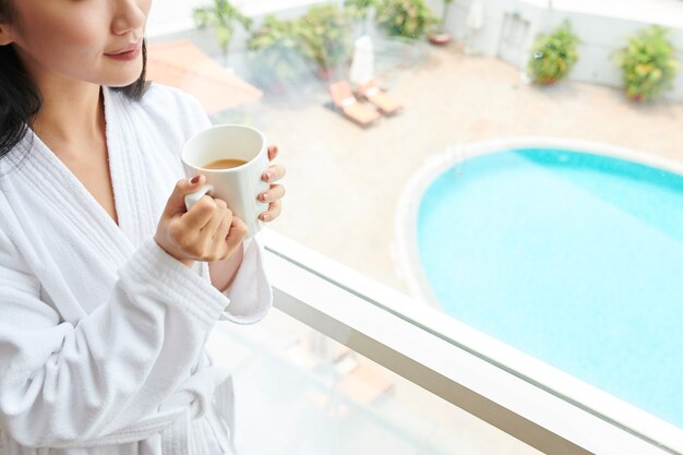 Bijgesneden afbeelding jonge vrouw in witte badjas die grote mok ochtendkoffie drinkt wanneer ze bij het raam van het hotel staat en naar buiten kijkt