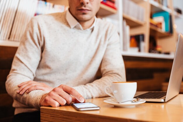 Bijgesneden afbeelding van een jonge aantrekkelijke man die in café zit terwijl hij een mobiele telefoon gebruikt en koffie drinkt.