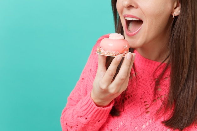 Foto bijgesneden afbeelding van jong meisje in gebreide roze trui houden in de hand eten taart geïsoleerd op blauwe turquoise muur achtergrond studio portret. mensen oprechte emoties, lifestyle concept. bespotten kopie ruimte.