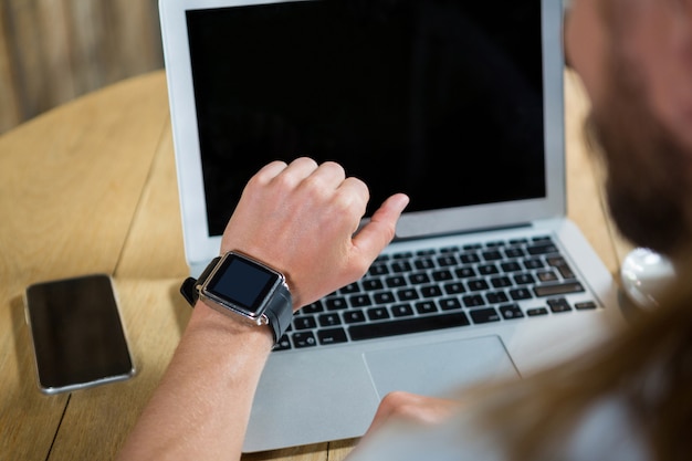 Bijgesneden afbeelding van jonge man met behulp van slimme horloge met laptop en mobiele telefoon op tafel in café