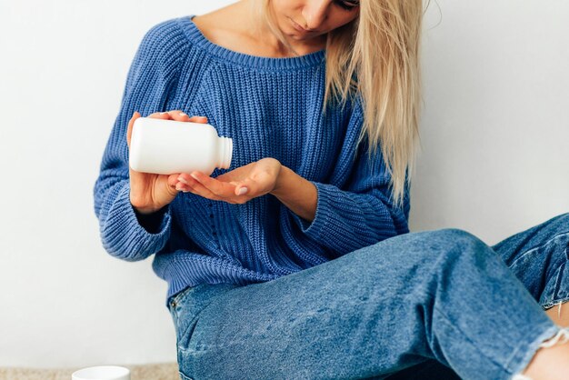 Bijgesneden afbeelding van jonge vrouw giet de pillen uit de fles Jonge blonde vrouw draagt blauwe gebreide trui die wat pillen in haar hand uit een witte fles giet Gezondheidszorg en geneeskunde