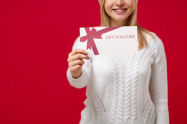 Bijgesneden afbeelding van jonge vrouw in witte trui hoed geïsoleerd op rode achtergrond studio portret. Gezonde mode levensstijl mensen emoties, koud seizoen concept. Mock up kopie ruimte. Cadeaubon vasthouden.