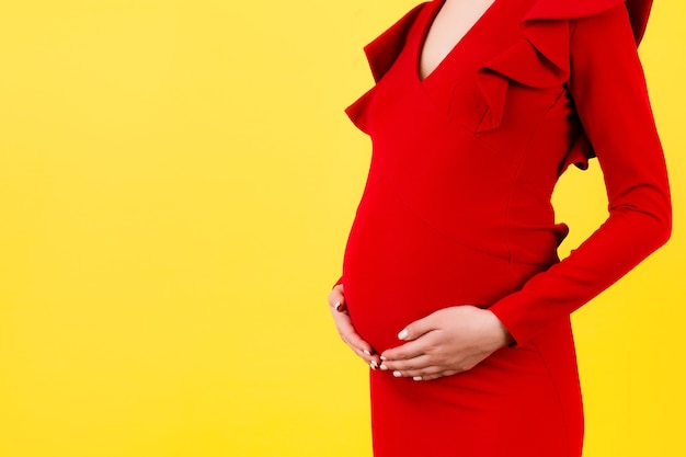 Bijgesneden afbeelding van zwangere vrouw die rode jurk draagt. Jonge moeder knuffelt haar buik en verwacht een baby op gele achtergrond. Ruimte kopiëren.