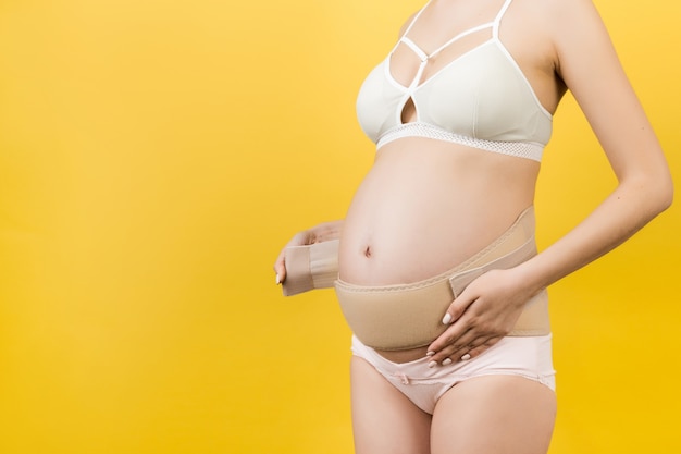 Bijgesneden afbeelding van zwangere vrouw in ondergoed die ondersteunend verband aanbrengt voor het verminderen van rugpijn op gele achtergrond met kopieerruimte. Orthopedisch buiksteunriemconcept.