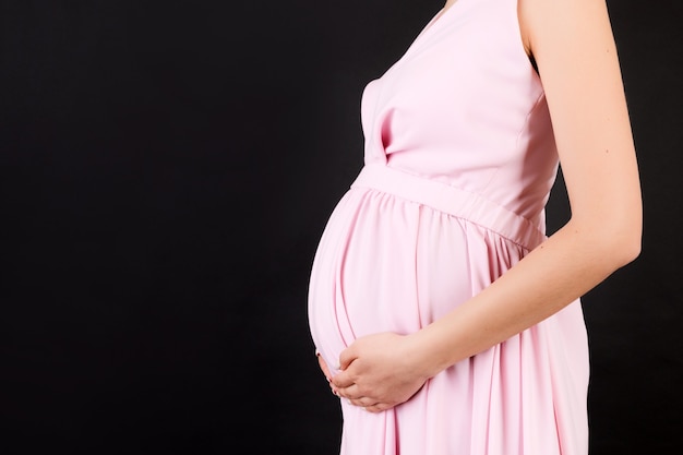 Bijgesneden afbeelding van zwangere vrouw in roze jurk omarmen haar grote buik op zwarte ondergrond