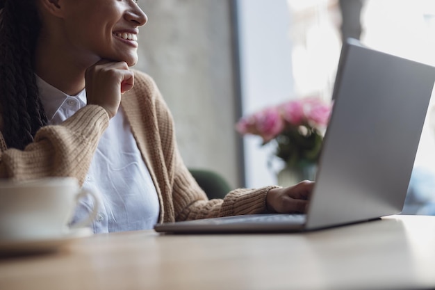 Foto bijgesneden beeld van een jonge afrikaanse vrouw glimlacht dromerig terwijl ze uit het raam naar de straat in de stad kijkt terwijl ze op afstand op een laptop werkt