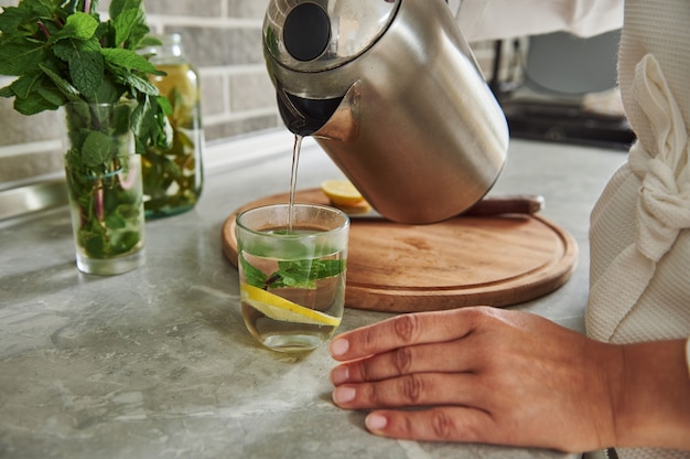 Foto bijgesneden beeld van vrouw in gewaad die water uit theepot giet in glas met muntblaadjes en schijfje citroen en gezond water bereidt met munt en citroen. detailopname.