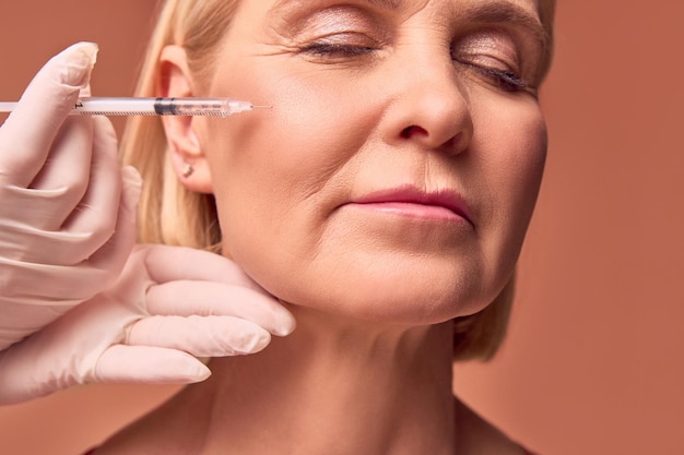 Foto bijgesneden close-up portret van een volwassen vrouw met gesloten ogen op een beige achtergrond handen in witte handschoenen en een medische jurk maken een injectie in het jukbeen