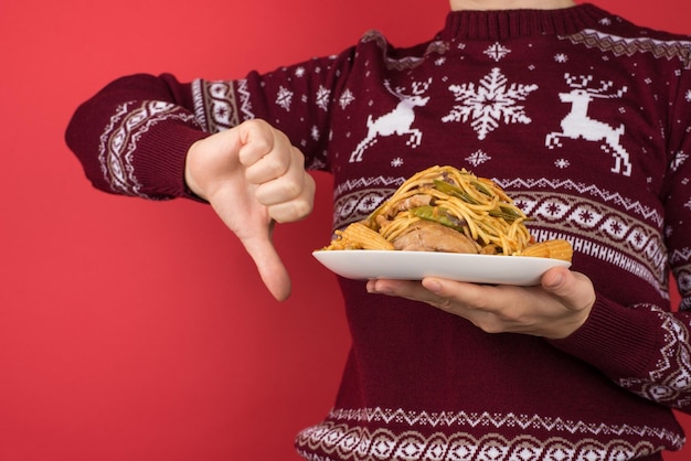 Bijgesneden close-upfoto van meisje in rode en witte kersttrui die een groot bord eten vasthoudt en duim naar beneden maakt op geïsoleerde rode achtergrond