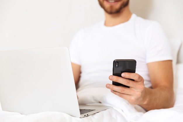 Bijgesneden een knappe jonge bebaarde man in bed thuis met behulp van laptop en mobiele telefoon.