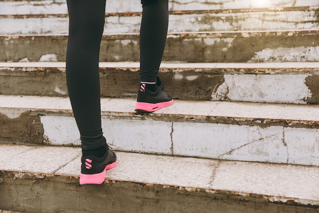 Bijgesneden foto close-up van vrouwelijke benen in sportkleding, zwarte en roze vrouwensneakers die sportoefeningen doen, buitenshuis op trappen klimmen