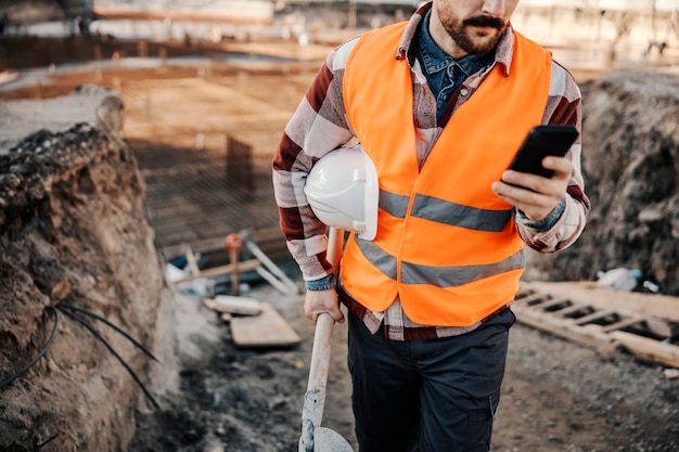 Bijgesneden foto van een bouwplaatsmedewerker die een telefoon gebruikt terwijl hij op de bouwplaats loopt