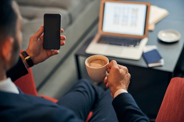 Bijgesneden foto van een man in een pak die zit met een kopje koffie en een laptop op de achtergrond en een moderne mobiele telefoon vasthoudt