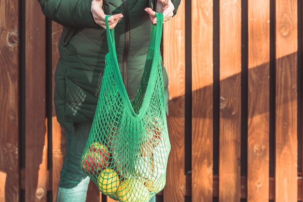 Bijgesneden foto van onherkenbare vrouw met blauwe gebreide mesh tas Outdoor foto gebreide tas vol met groenten en fruit in handen van de vrouw houten planken op een achtergrond kopie ruimte