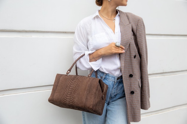 Bijgesneden foto van stijlvolle vrouw in wit overhemd en oversized jas met bruine tas tegen muur in straat