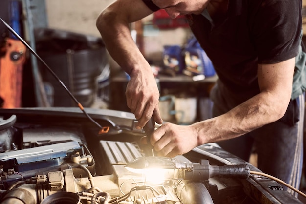 Bijgesneden handen van onherkenbare monteur close-up reparateur met schroevendraaier en glanzende lamp die onder de motorkap van de auto werkt