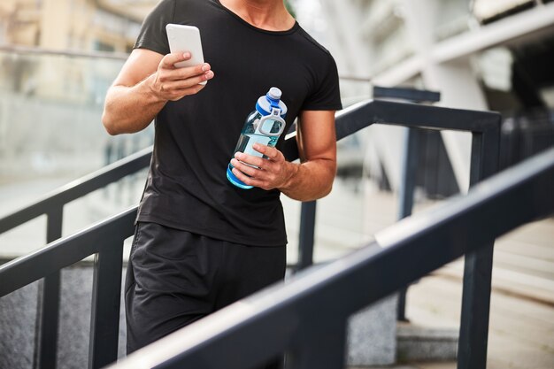 Bijgesneden hoofd van atletische man met water en berichten op smartphonetraining in de stad