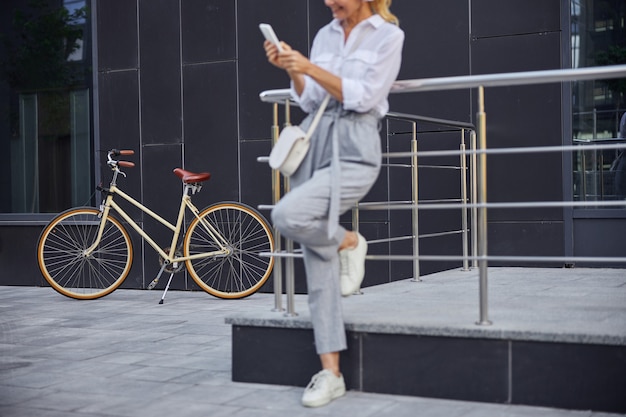 Bijgesneden hoofdportret van retro stadsfiets waar een vrouw in zakelijke vrijetijdskleding ervoor staat tijdens het gebruik van een smartphone