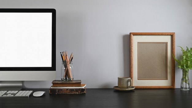 Bijgesneden opname van computer met wit leeg scherm, muis en toetsenbord zetten op zwarte houten tafel naast potlood houder, oude boek, koffiekopje, fotolijst en potplant. vintage bureau.