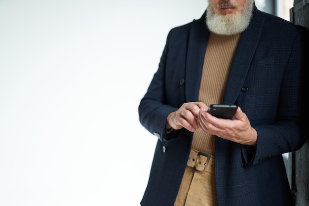 Bijgesneden opname van drukke volwassen man met baard in zakelijke vrijetijdskleding met smartphone leunend