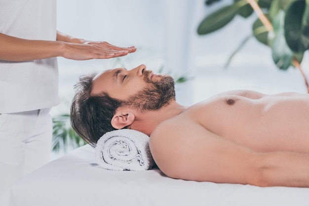 Bijgesneden opname van een bebaarde man met gesloten ogen die liggen en reiki-therapie boven het hoofd ontvangen