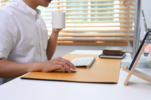 Bijgesneden opname van een jonge man die een koffiekopje vasthoudt en online werkt op het kantoor aan huis.