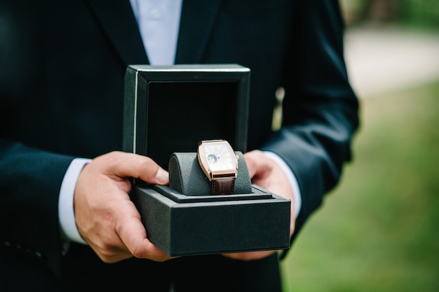 Foto bijgesneden opname van een man die een elegant pak draagt en een polshorloge vasthoudt. mensenhand met elegant duur luxehorloge met leerriem op een aardachtergrond.