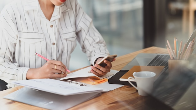 Bijgesneden opname van een vrouw in gestreept overhemd met een smartphone in de hand en schrijven / nemen notitie zittend aan het houten bureau.