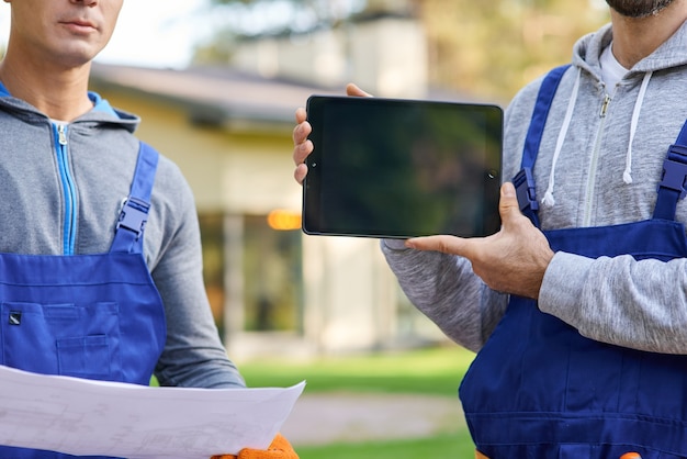 Bijgesneden opname van jonge mannelijke bouwer die scherm van digitale tablet-pc toont met collega die blauwdruk op de achtergrond vasthoudt, werkend op de bouwplaats van een huisje