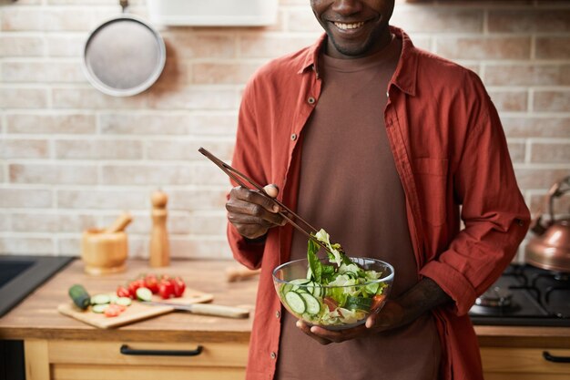 Bijgesneden opname van lachende zwarte man die salade in glazen kom mengt terwijl hij geniet van een gezonde maaltijd in een gezellige kopieerruimte in de keuken