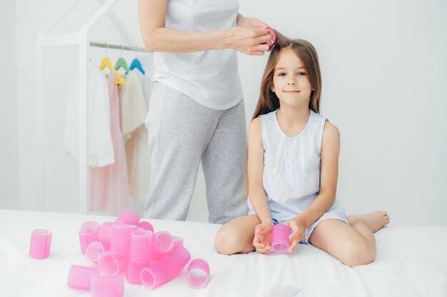 Bijgesneden opname van onherkenbare moeder windt krulspelden naar haar mooie dochter die een mooi kapsel gaat maken heeft een aangename uitstraling en lang haar Schattig lief klein meisje wil krullen hebben