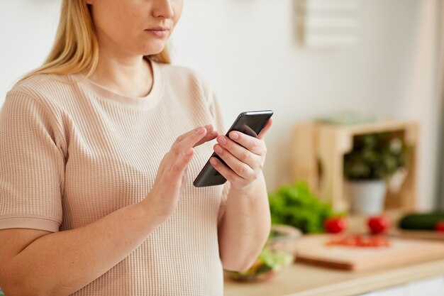 Bijgesneden portret van een volwassen vrouw met behulp van smartphone en tekstbericht typen terwijl je in gezellige keuken interieur, kopie ruimte