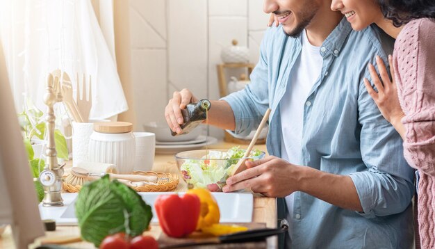 Bijgesneden schot van jonge arabische familie die samen groentesalade in de keuken bereidt, onherkenbare echtgenoten uit het midden-oosten die genieten van het koken van gezond voedsel, man die olijfolie in de kom giet, panorama