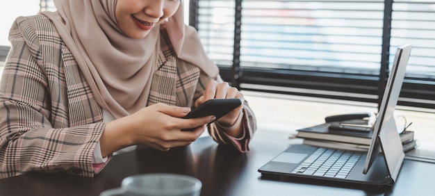 Bijgesneden schot van moslim zakenvrouw in bruine hijab en vrijetijdskleding zitten en smartphone gebruiken.