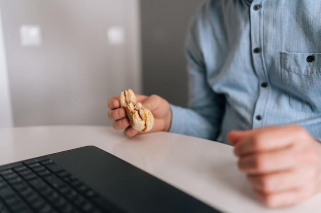 Bijgesneden schot van onherkenbare volwassen oude zakenman met rimpelhanden hamburger met rundvlees eten van fastfoodrestaurant zittend aan tafel online werken met laptopcomputer in kantoor aan huis