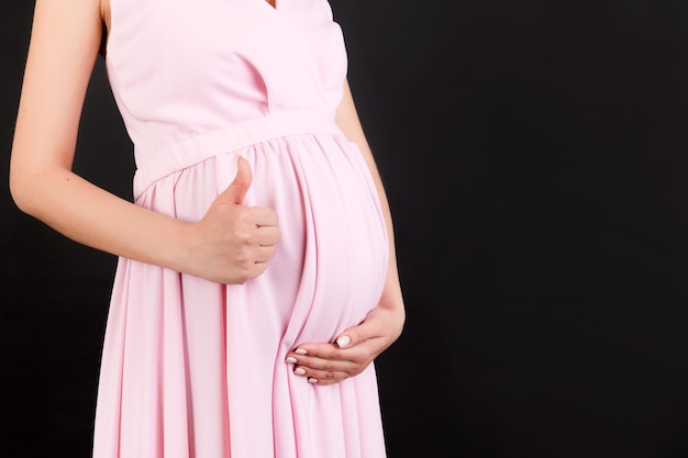 Bijgesneden schot van positieve zwangere vrouw in roze jurk met duim omhoog cool gebaar over haar babybuil op zwarte achtergrond. Gelukkig moederschap. Ruimte kopiëren.