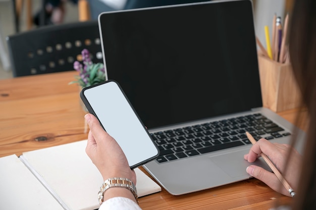 Bijgesneden schot van vrouw hand met leeg scherm smartphone tijdens het werken op laptopcomputer.