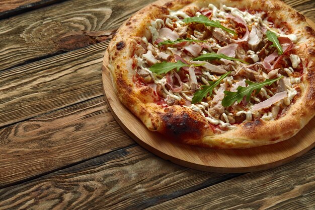 Bijgesneden shot van Napolitaanse pizza met mozzarella kaas, spek, ham, tomatensaus, kip, kruiden en verse rucola. Italiaanse pizza op houten tafel achtergrond