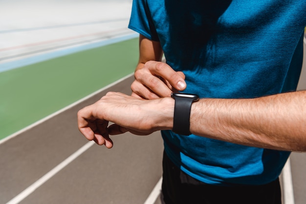 bijgesneden weergave van jonge sportman met behulp van smartwatch op atletiekbaan op zonnige dag