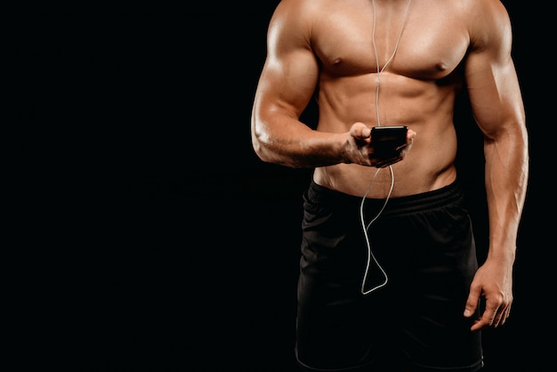 Bijgesneden weergave van sexy bodybuilder met gespierde romp luisteren muziek met koptelefoon op smartphone geïsoleerd op zwart