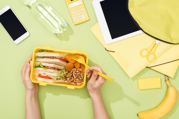 Bijgesneden weergave van vrouwenhanden met plastic gebruiksvoorwerpen over lunchbox met voedsel in de buurt van digitale rugzak