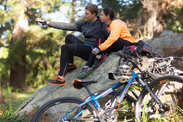 Biker couple sitting on rock pointing at distance in forest