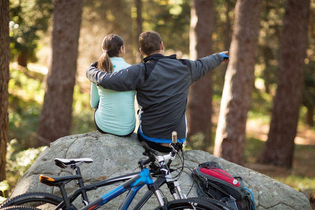 Biker couple sitting on rock pointing at distance in forest