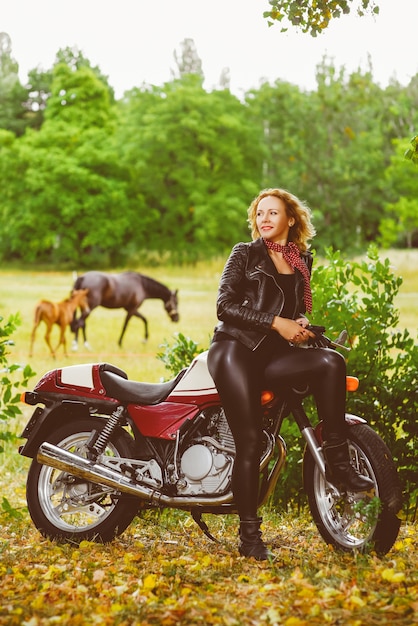 Biker girl in a leather jacket on a motorcycle against the background of horses.